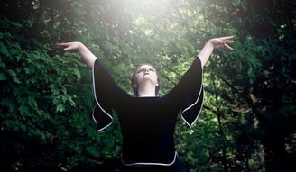 a woman with her arms outstretched up and she is looking up to the sun in the forest