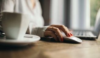 a woman at a laptop with hand on a mouse and a coffee mug on table