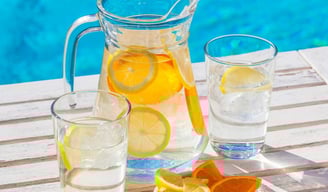 a jug and two glasses with water, ice and lemon and orange slices with a swimming pool in the backgr