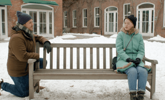 A young man leans on one end of a park bench, while a young woman sits eyes closed on the other end