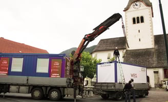 Kran bei der Aufstellung des Ausstellungscontainers "Aus weg los!" auf einem Dorfplatz.