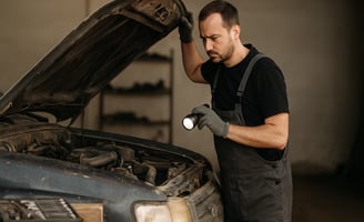 Mecânico levanta o capô de carro desgastado e inspeciona o motor com lanterna em oficina simples.