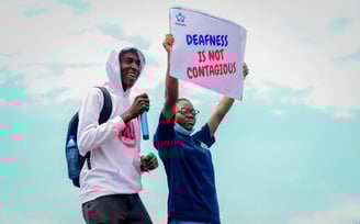 A man in a white hoodie, standing next to a woman who is holding up a banner that says "Deafness is not contagious"