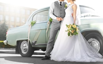 Image of a couple in wedding dress and formal suit standing beside an automobile.