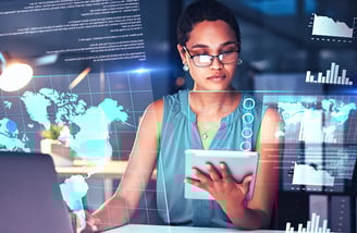 A female leader with glasses holds a tablet and types on a laptop at her desk, surrounded by holographic maps and graphs.