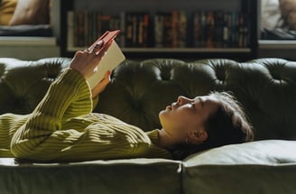 Uma jovem tranquilamente em um sofá, lendo um livro de artes visuais.