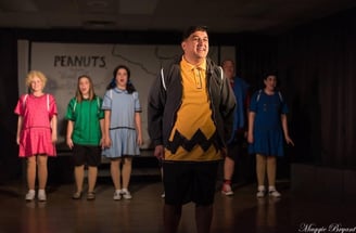 an actor playing Charlie Brown in a yellow sweater and black shorts standing in front of a group of