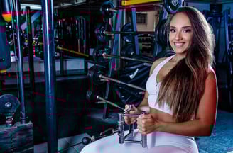 atleta de cabelos longos vestindo conjunto fitness branco e sorrindo no aparelho da academia