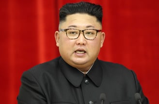 Kim Jong-un in a suit and glasses is standing Infront of a microphone