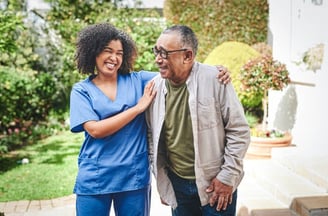 A nurse stands beside an elderly man outside, both smiling and engaged in conversation in a sunny ou