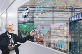 a woman in a black jacket and glasses is pointing at a transparent display of cars