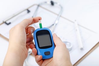 Hand holding a blood glucose meter measuring blood sugar, the background is a stethoscope and chart