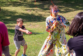 Summer Camp children shooting paint at the teacher in a white trenchcoat.