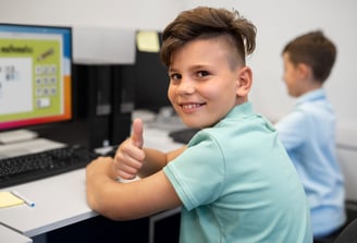 Smiling student giving thumbs up while using a computer in a classroom