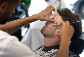 man receiving a shave with a cut throat razor by a barber while sitting in a chain in a barbershop