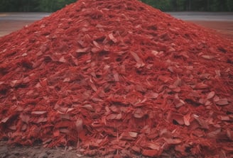 A collection of small, irregularly shaped mulch fragments in shades of red and pink mixed with some twigs and a small green plant in the lower left corner.