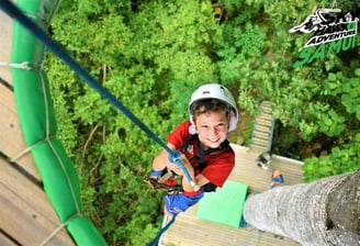 A person is zip-lining across a lush, green landscape, surrounded by dense foliage and rocky outcrop