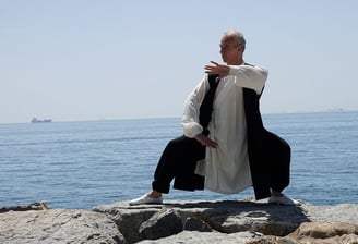 Esat Ataç holding a Tai Ji posture on seaside rocks, wearing traditional Wudang clothing.