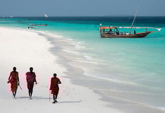 three people walking on a beach with boats in the background