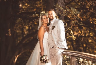 Brautpaar in eleganter Hochzeitskleidung auf einer Holzbrücke umgeben von Bäumen