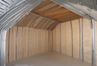 A house is under construction with visible wooden framing and scaffolding around the roof area. The building is enclosed by a temporary metal fence, and there are materials and construction tools placed around the site. The roofing is partially complete, made of dark grey tiles, and the surrounding area includes grass and a paved sidewalk.