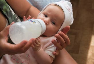 A baby wearing a striped hat and green sweater is being breastfed. The scene is intimate and tender,