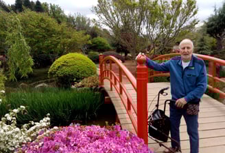 Older man standing at the foot of a beautiful red bridge in an ornamental garden with his walker