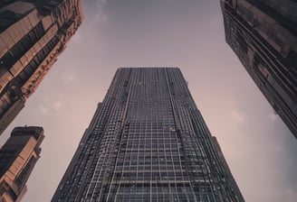 A modern skyscraper with the PwC logo is partially obscured by out-of-focus autumn leaves in the foreground. The building is set against a backdrop of a bright, cloudy sky.