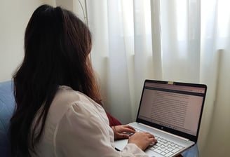 Woman typing on a laptop with a document open, sitting near a window with white curtains.