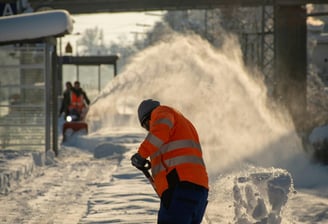 A crew of men in high visibility clothing snowblow and shovel snow