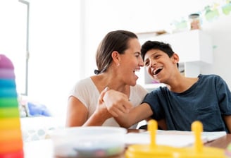 A woman and a boy share a joyful moment, laughing together at a table.