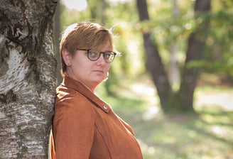 Femme avec lunettes et veste marron adossée à un arbre en forêt