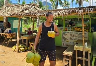 a man holding a drink in front of a hut with coconut trees