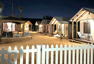row of hemp town cabins well lit in dusk behind white picket fence