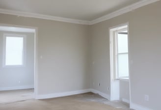 A partially constructed or renovated interior space featuring exposed brick walls and wooden ceilings. A white door is visible in the background next to a red painted wall. A plastic sheet covers furniture on the right side, and there are construction materials scattered across the floor.