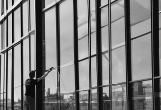 Two window cleaners are suspended by ropes, working on cleaning the glass windows of a tall building. Each cleaner is equipped with tools and a bucket, skillfully moving along the facade with safety harnesses. The building features reflective blue-tinted glass panels, creating a striking geometric pattern.