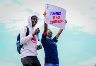 A man in a white hoodie, standing next to a woman who is holding up a banner that says "Deafness is not contagious"