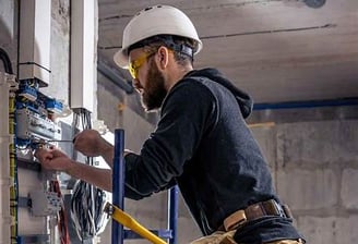 Alt="Commercial electrician installing wiring in a business facility, wearing hard hat and safety."