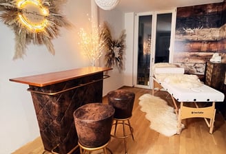 Auf dem Bild ist eine braune Bar, mit zwei braunen Barhockern, Massageliege mit weißem Leder, auf der ein weißes Lammfell