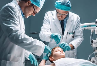 A dentist is performing a procedure on a patient reclining in a dental chair. The dentist is wearing a protective cap, mask, and gloves while using dental tools. The room features modern decor with geometric patterns on the wall, and there is a dental light overhead. Nearby, there is a table with dental instruments and a monitor on the wall.