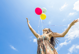 Mujer joven en un espacio abierto que muestra un cielo azul, lanzando tres globos al aire, feliz.