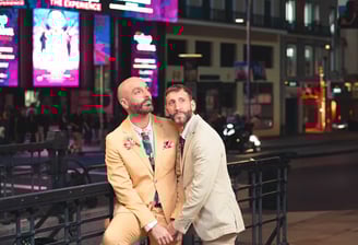 Pareja gay feliz tocándose la frente y sonriendo en una calle metropolitana de España, luz natural y momento espontáneo