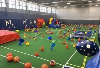 A large indoor sports facility featuring a green playing surface, similar to a bowling green. The ceiling is lined with numerous fluorescent lights, giving the space a bright yet serene atmosphere. Red chairs are arranged along the walls, and a scoreboard can be seen in the distance. A person is actively engaged in the game, bending forward as they release a ball, while inset a few spectators or participants are visible on the left side of the space.