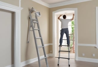People are working on the interior renovation of a room that appears to be under construction. A worker on a scaffold is painting the ceiling, while others are seated or discussing the progress. The room has smooth, gray walls with arched recesses and a modern design vibe. There are tools and materials scattered around, including a ladder, paint cans, and some rocks or construction debris on the floor. Overhead, stylish pendant lights provide illumination as natural light streams in from windows.
