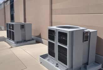 A series of air conditioning units are mounted on a metal structure outside a building with white paneling. Two large windows are visible behind the units, revealing the indoors with fluorescent lighting.