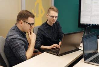 Dois adultos em frente a um computador, sendo que um deles está ensinando o outro como operar