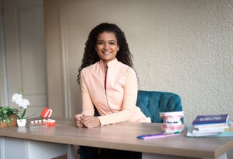 Mujer joven con cabello rizado sentada en un escritorio, sonriendo y rodeada de libros, modelos dentales y plantas en una ofi