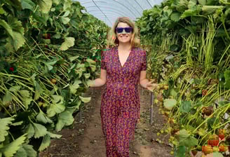 a woman in a navy blue jumpsuit with red strawberries and sunglasses standing by strawberry plants