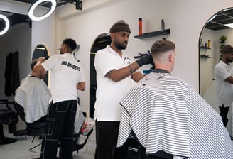 two barbers tending to cutting the hair of two customers while they are sitting in the chair