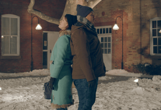 A young woman and man in winter clothing lean their backs against each other in a snowy courtyard